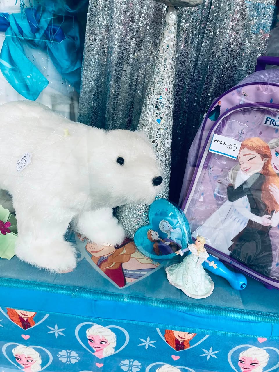 Frozen frenzy at AAPS Op Shop Cheltenham, with children’s Elsa and Anna costumes, dolls, toys, glitter shoes and wintery clothing in store. Visible prices include a Frozen backpack for $5, a polar-bear soft toy for $8, an upcycled doll for $10, and a white crystal dress in size 12–14 for $45.