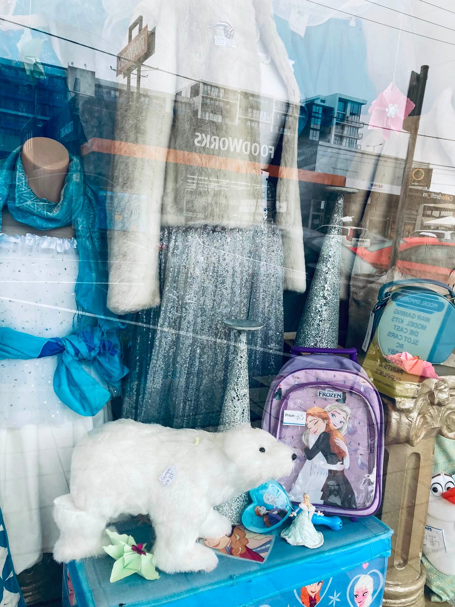 Frozen frenzy at AAPS Op Shop Cheltenham, with children’s Elsa and Anna costumes, dolls, toys, glitter shoes and wintery clothing in store. Visible prices include a Frozen backpack for $5, a polar-bear soft toy for $8, an upcycled doll for $10, and a white crystal dress in size 12–14 for $45.