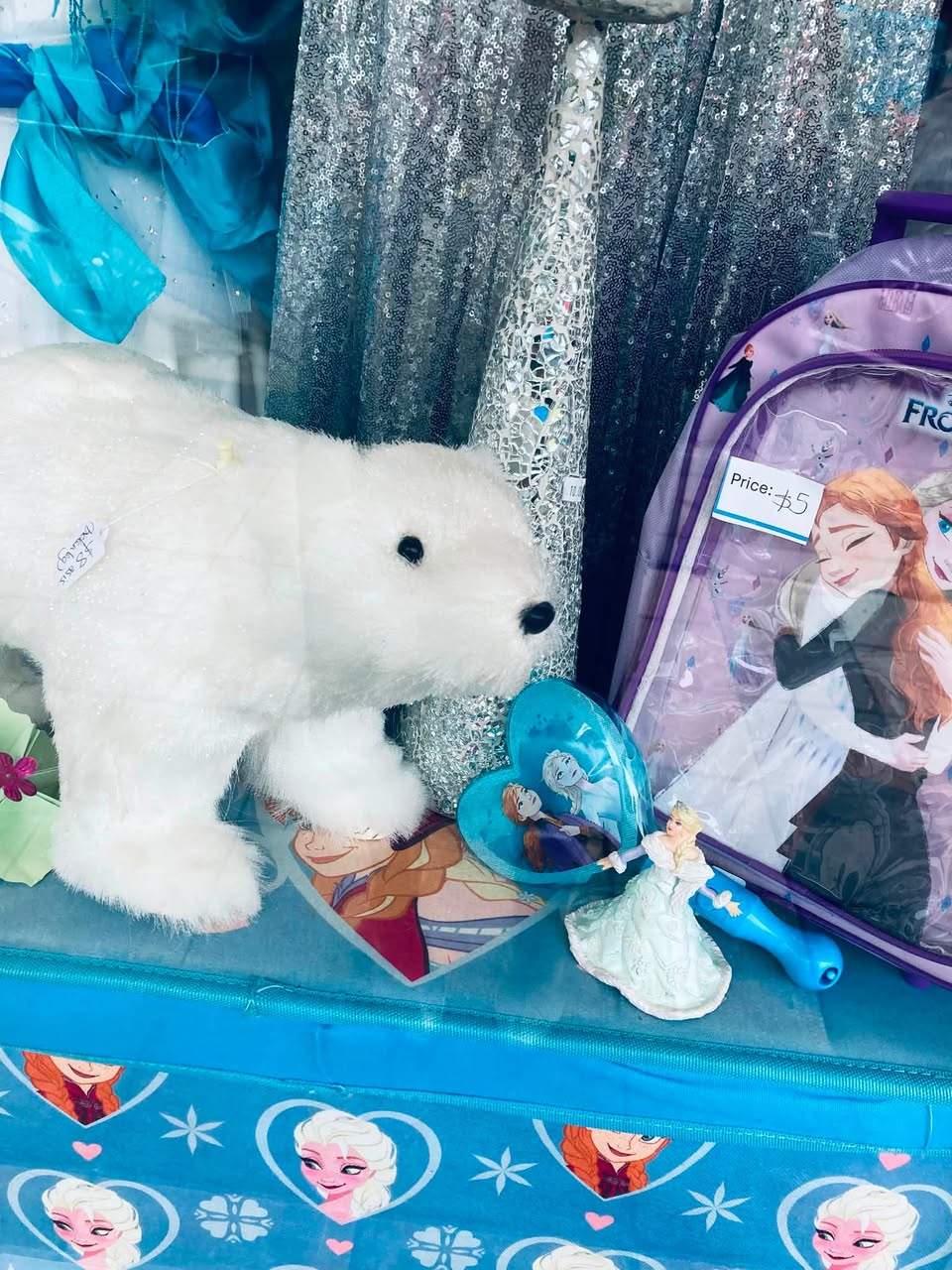 Frozen frenzy at AAPS Op Shop Cheltenham, with children’s Elsa and Anna costumes, dolls, toys, glitter shoes and wintery clothing in store. Visible prices include a Frozen backpack for $5, a polar-bear soft toy for $8, an upcycled doll for $10, and a white crystal dress in size 12–14 for $45.