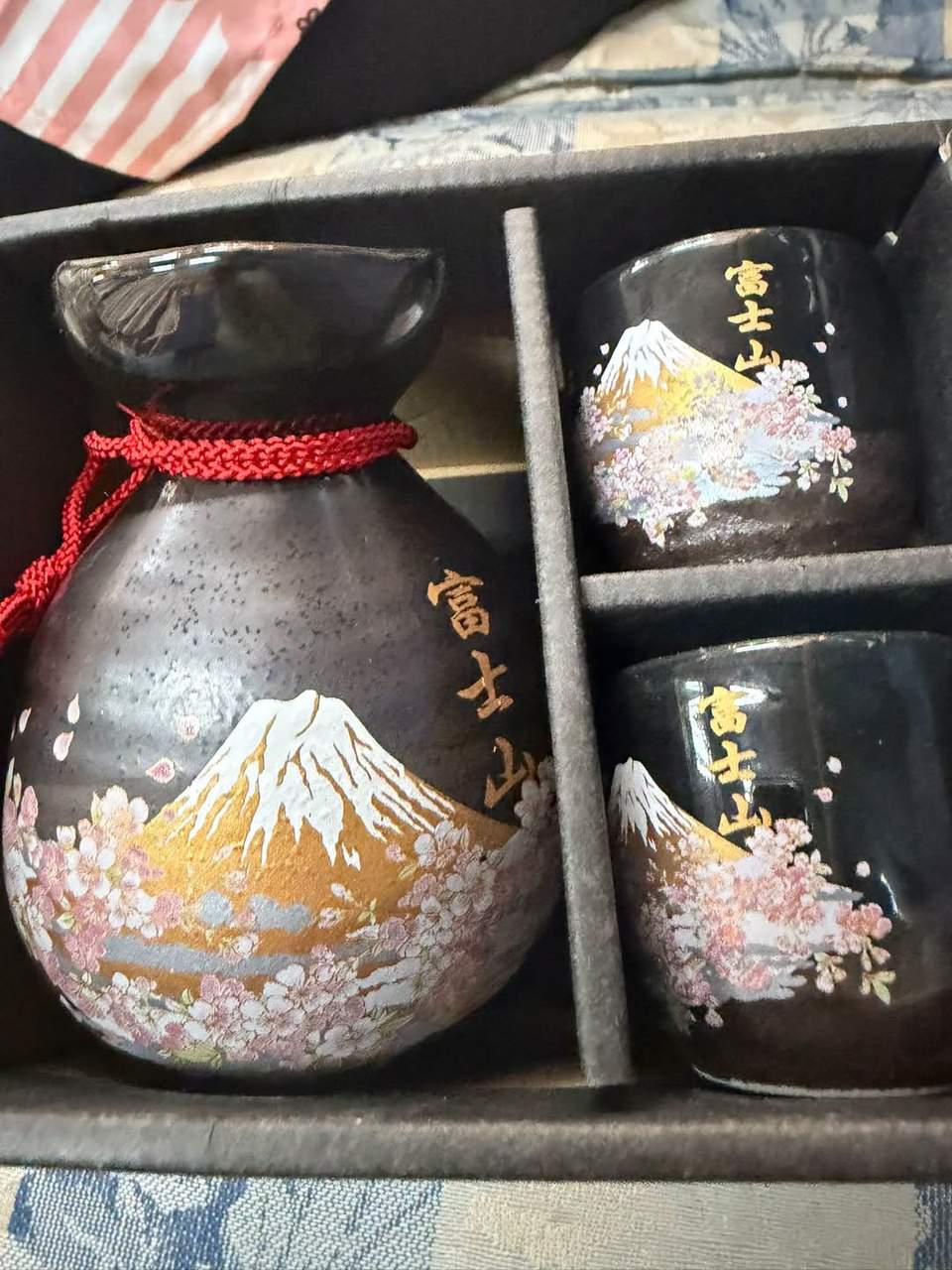 The image shows a black ceramic Japanese sake set featuring a painted Mount Fuji and cherry blossoms design, complete with a tokkuri (flask) and two matching ochoko (cups) in a presentation box.